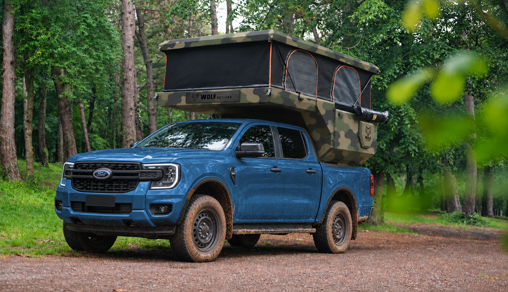 Front three-quarter view of a blue Ford Ranger with an Army camouflage WOLF CAMPER pickup camper, pop-up roof raised among the trees.