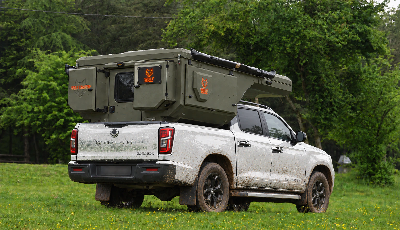 Rear three-quarter view of a white KGM Musso with a tactical olive WOLF CAMPER pickup camper in a clearing.