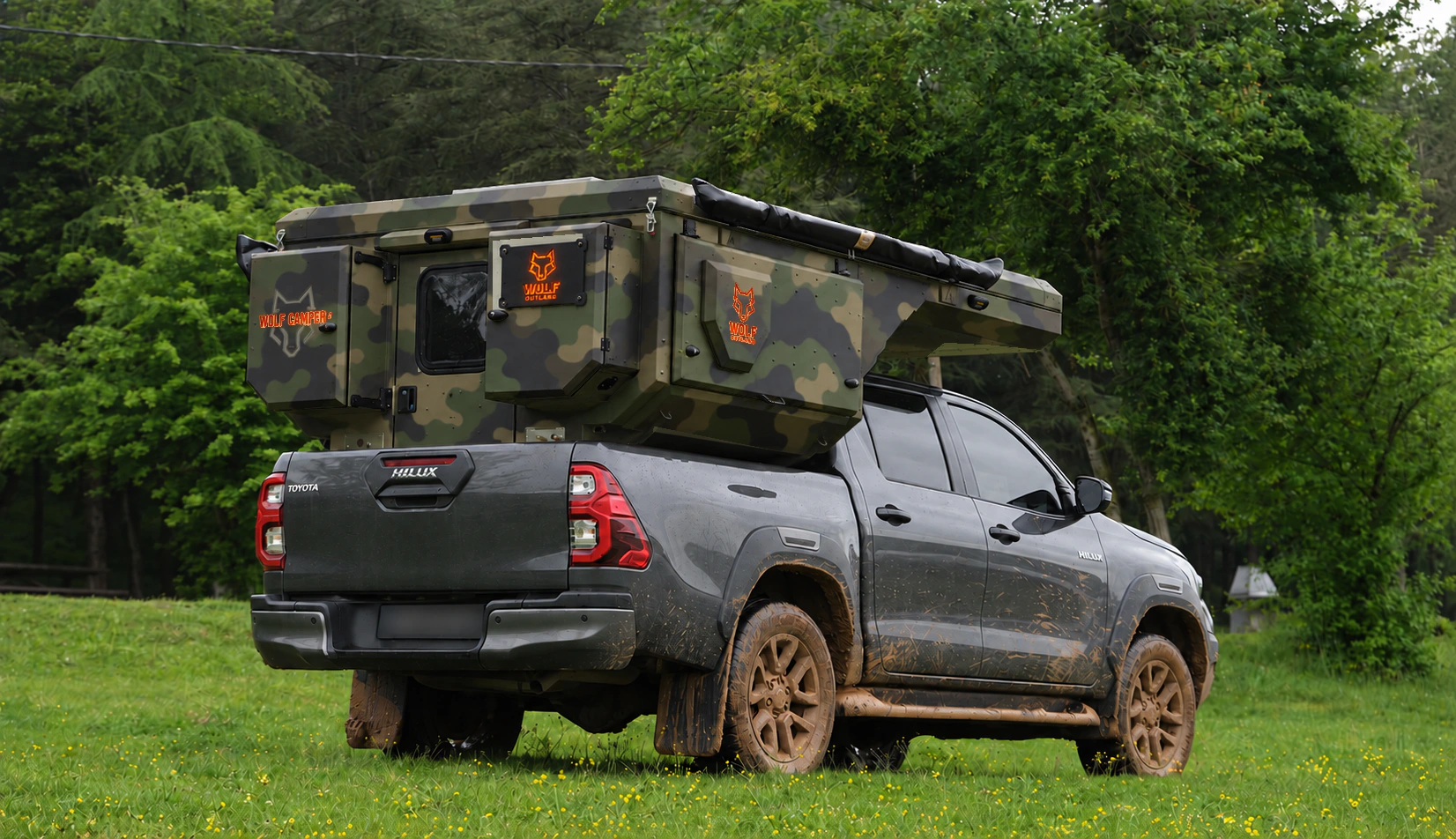 Toyota Hilux in anthracite grey with WOLF CAMPER in Army camouflage, solid cabinets — Rear right three-quarter, Roof closed, Orange backlighting.