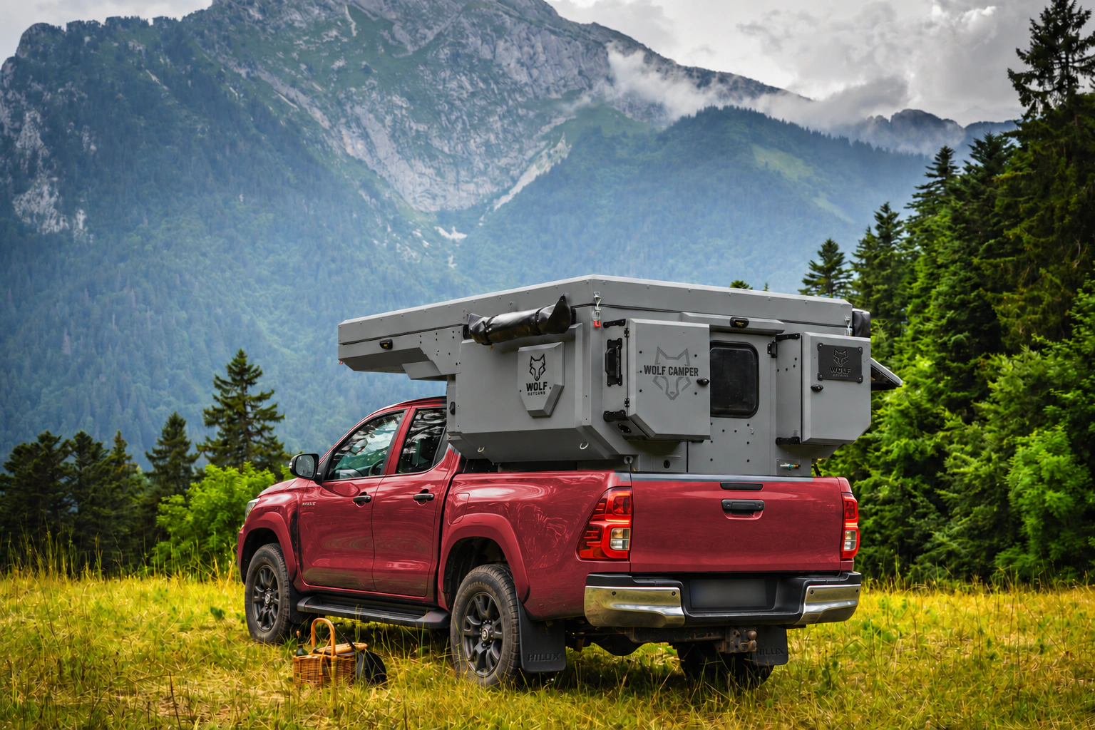 Rear three-quarter view of a red Toyota Hilux with a mineral grey WOLF CAMPER pickup camper in a meadow facing the mountains.