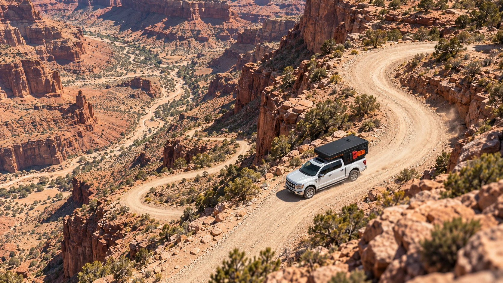 High drone view of a silver Ford F-150 Lariat with a matt black WOLF ADVENTURE pickup camper on a winding track through a red canyon.